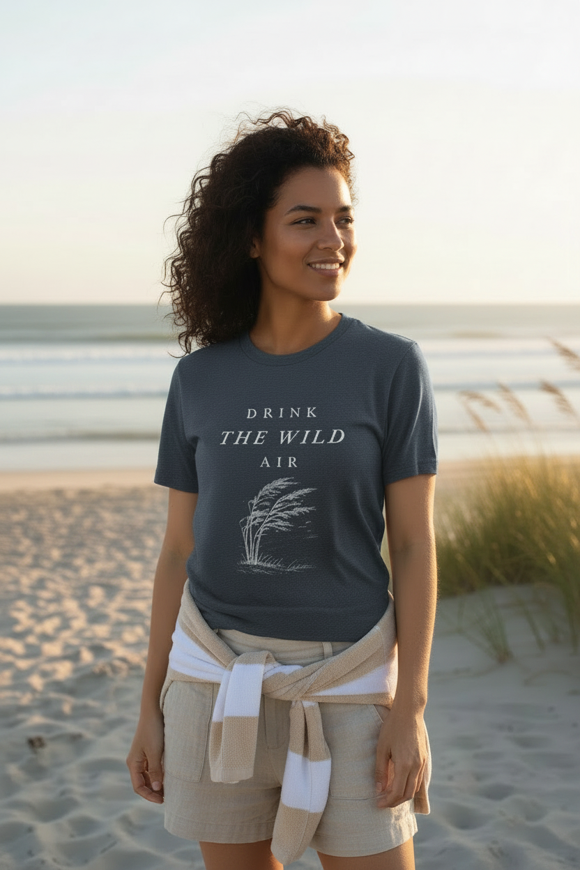 Woman standing on a beach wearing a t-shirt with 'Drink the Wild Air' text in heather blue