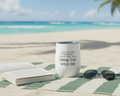 White tumbler with Emerson quote on a beach with an open book and sunglasses.
