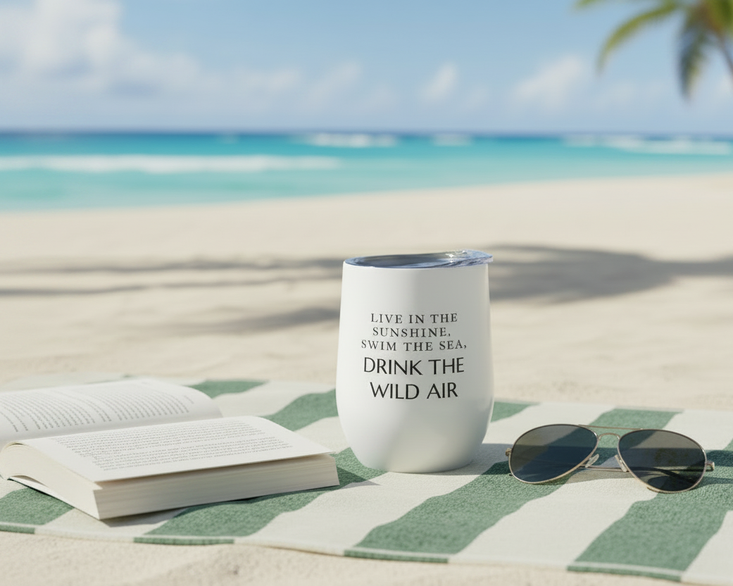 White tumbler with Emerson quote on a beach with an open book and sunglasses.