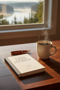 Book titled 'First Night Journal' with a steaming cup of coffee on a wooden table by a window with a view of trees and water.