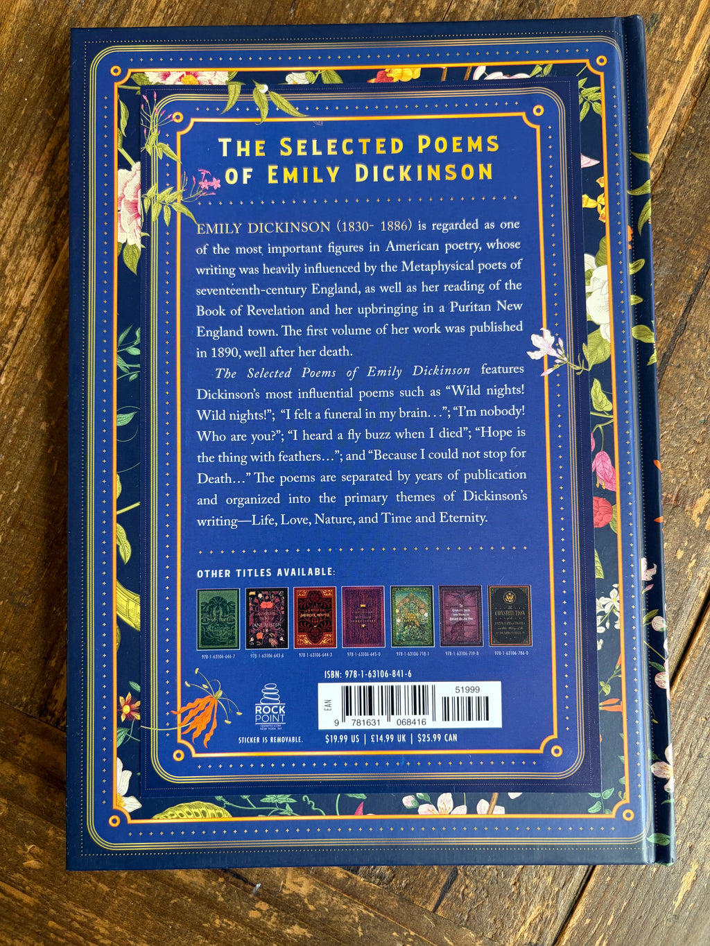 The back cover of the book 'The Selected Poems of Emily Dickinson' by Rock & Roll Books, displayed flat on a wooden surface. The cover features a dark blue background with gold borders, white text, and illustrations of birds and flowers.