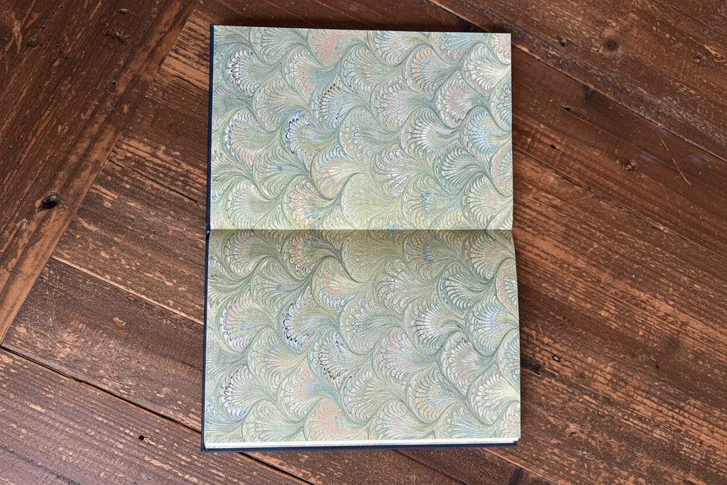 An open book or journal showing two pages with a marbled paper pattern in muted green, beige, and soft pink, held open by a hand on a rustic wooden table.