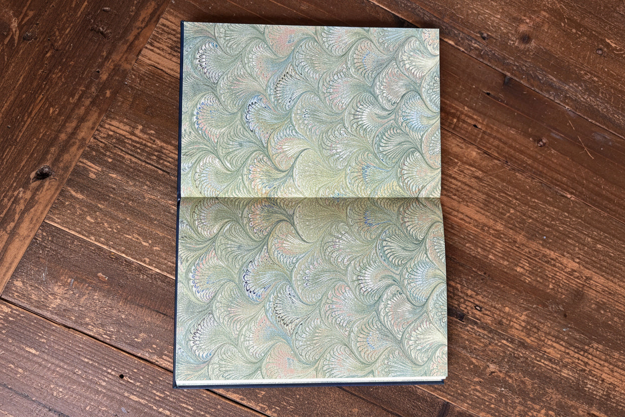 An open book or journal showing two pages with a marbled paper pattern in muted green, beige, and soft pink, held open by a hand on a rustic wooden table.