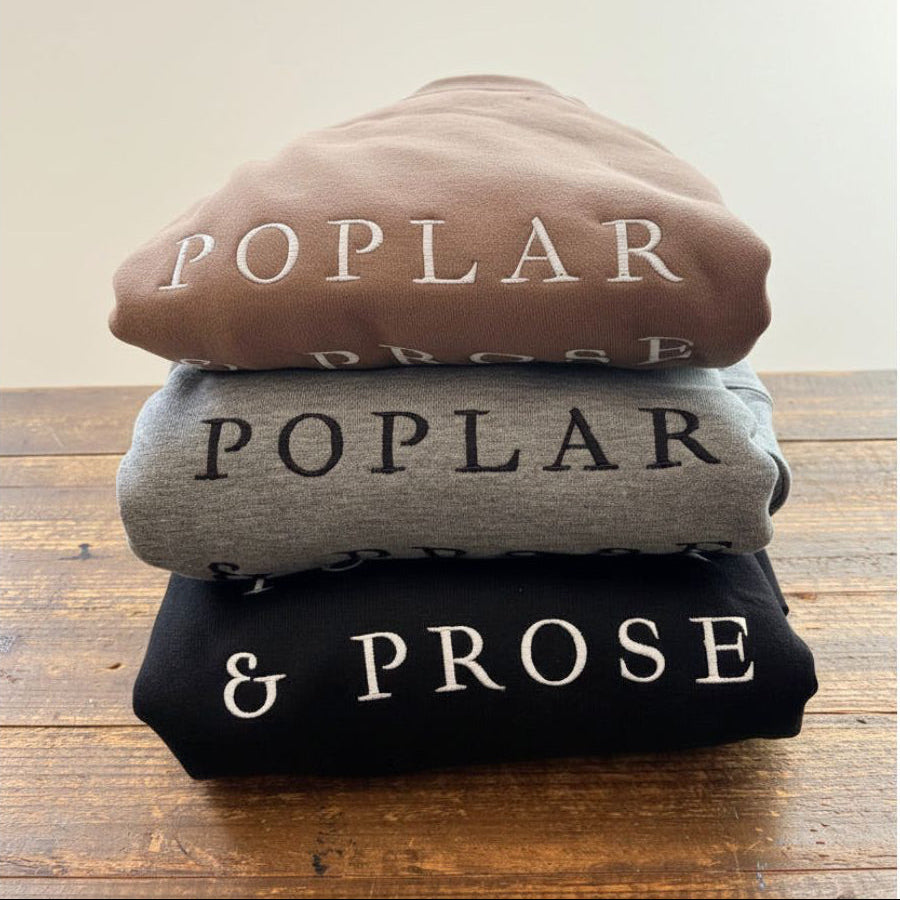 Stack of folded cotton sweatshirts with 'POPLAR & PROSE' branding on a wooden surface.