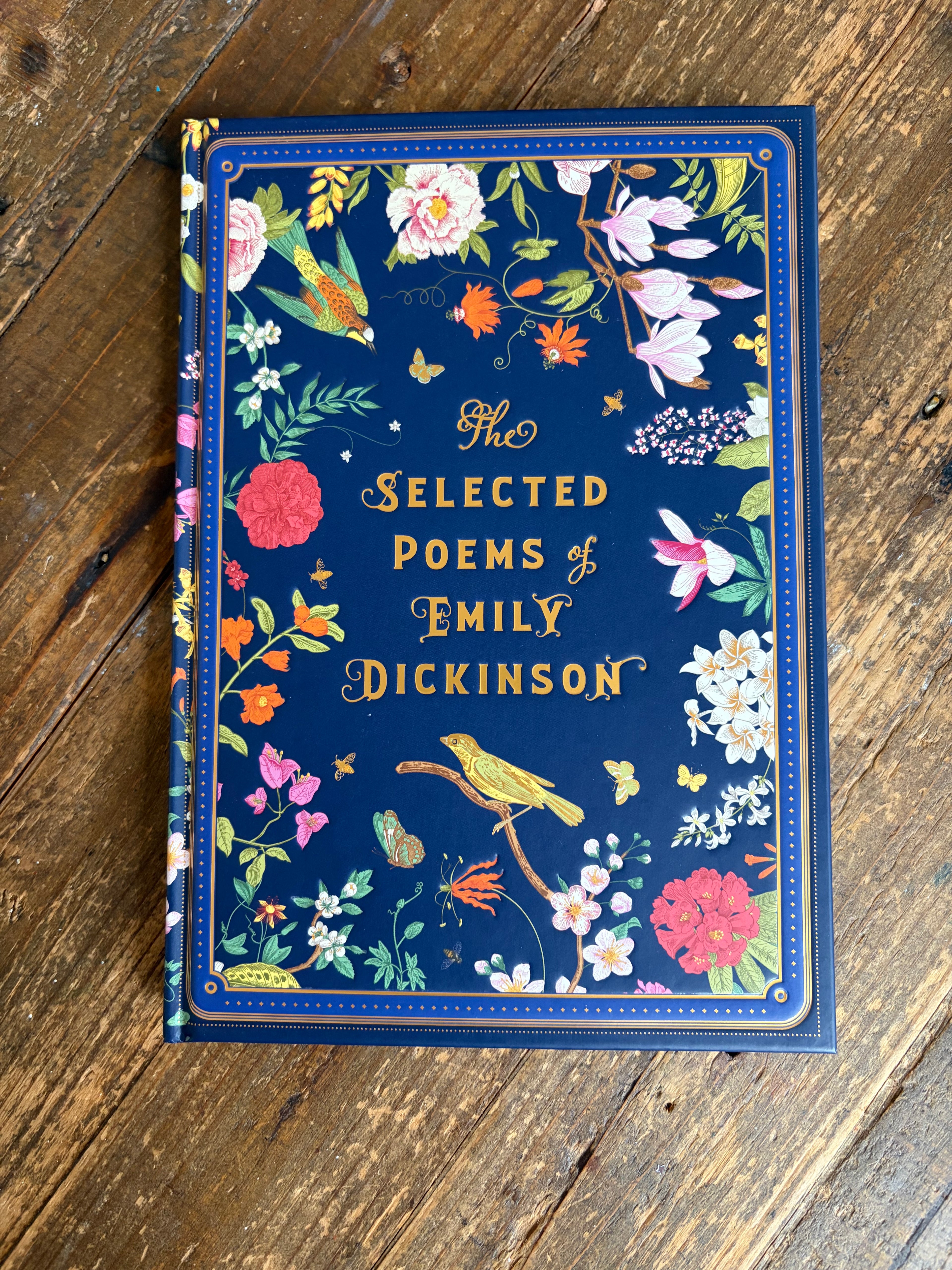 The Selected Poems of Emily Dickinson hardcover book with a navy blue floral cover, placed on a rustic wooden surface.