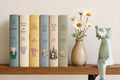 Six illustrated children's book spines stand on a wooden shelf beside a beige vase with white daisies and a pale blue elephant figurine. The books include titles such as The Story of Babar,  If Animals Know Good Night, Little Elliott Big City, and The Elephant in the Garden.