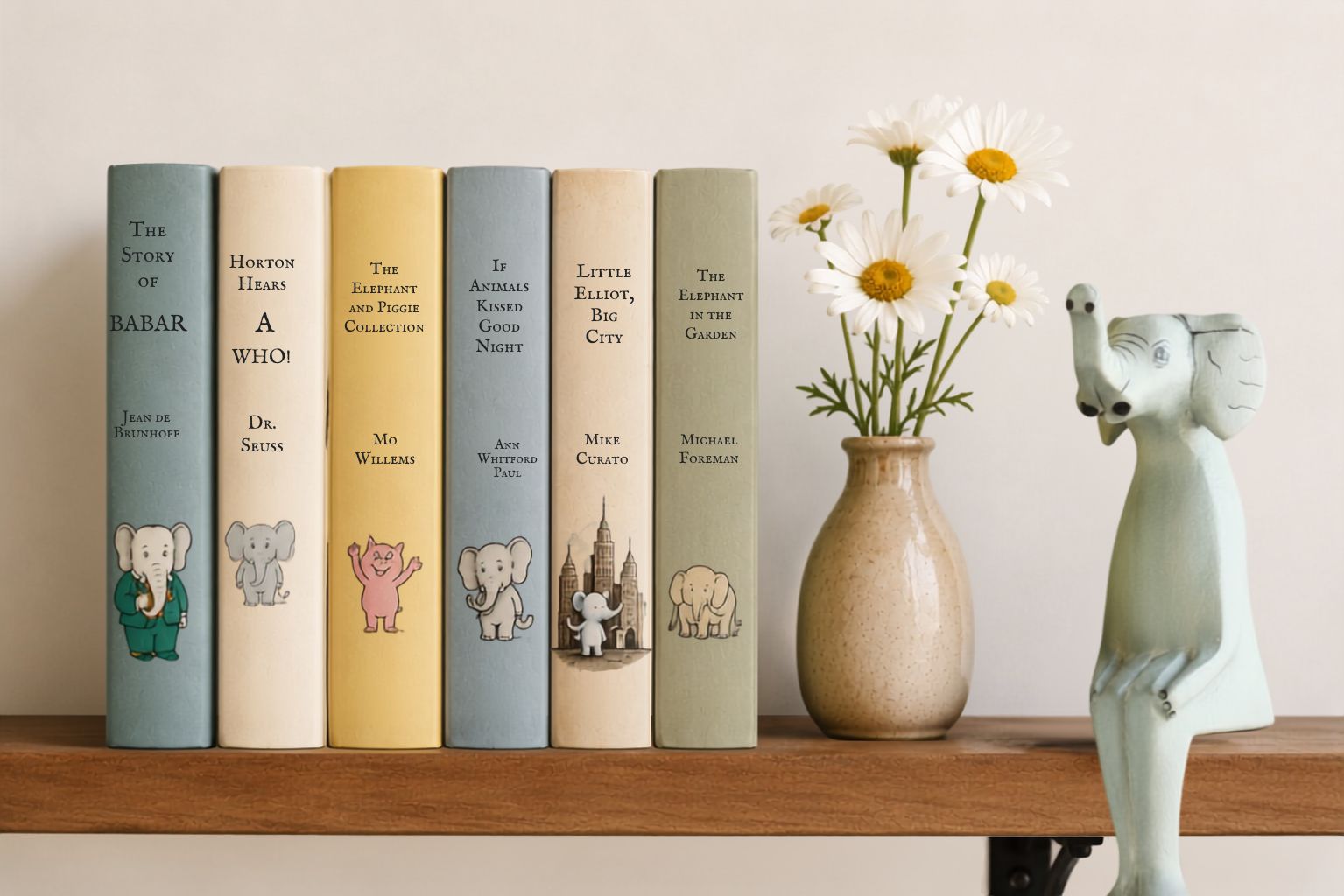 Six illustrated children's book spines stand on a wooden shelf beside a beige vase with white daisies and a pale blue elephant figurine. The books include titles such as The Story of Babar,  If Animals Know Good Night, Little Elliott Big City, and The Elephant in the Garden.