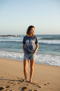 Woman standing on a beach with ocean waves in the background wearing the Grove organic cotton tee in blue