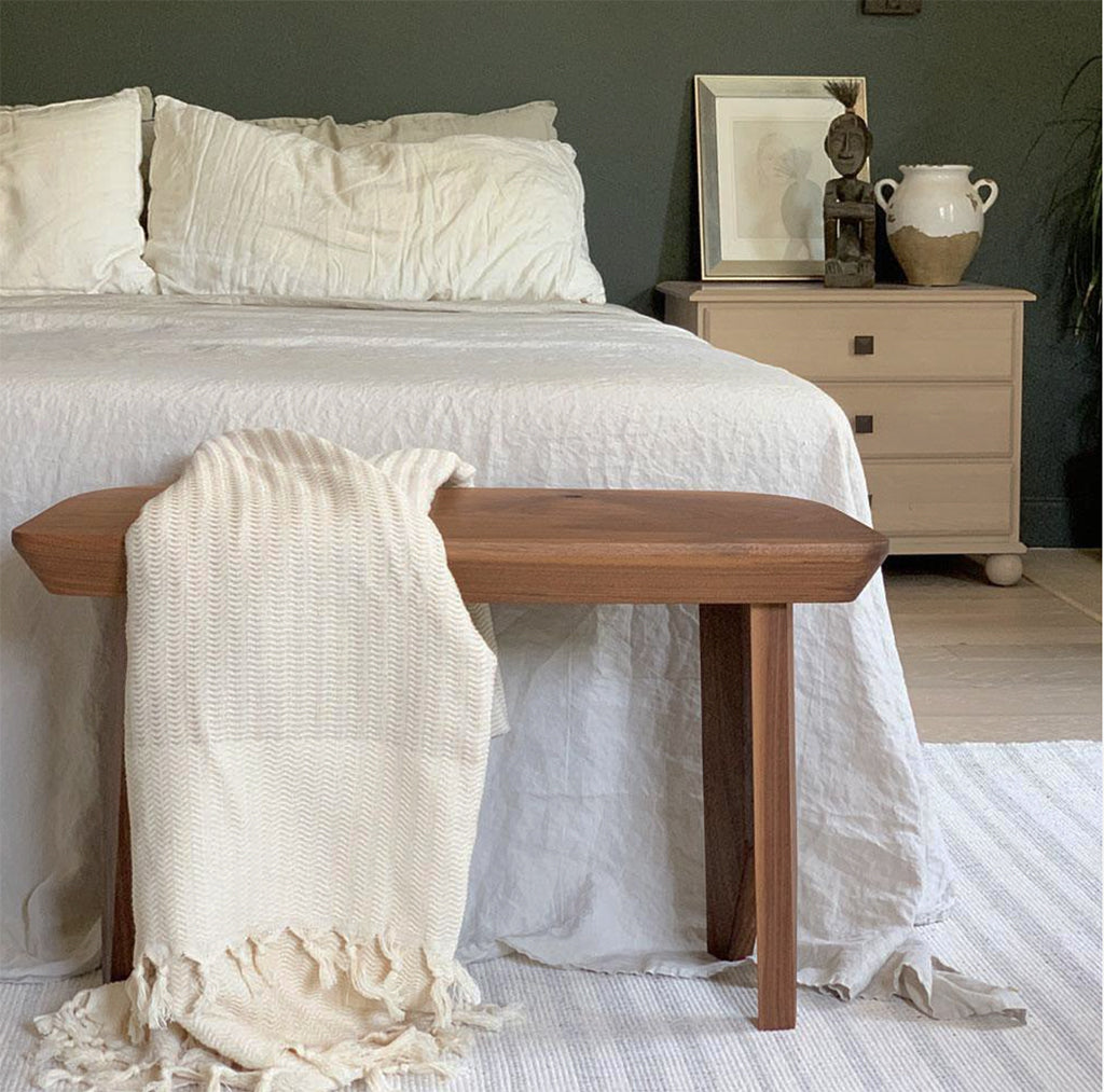 Wooden bench with a neutral blanket in front of a bed in a bedroom setting.
