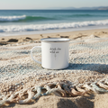 White enamel mug with 'drink the wild air' text on a sandy beach with ocean waves in the background