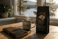 Steaming mug of coffee, leather notebook, and coffee bag on a wooden table with a window view of trees and water.