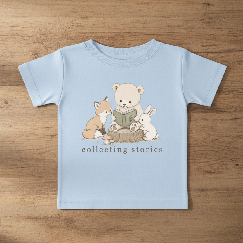 A light blue baby t-shirt laid flat on a wooden surface, featuring a cute graphic of a teddy bear, fox, and rabbit reading a book, with the text 'collecting stories' printed below.
