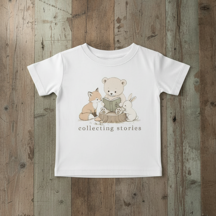 A white short-sleeve baby t-shirt with a cute graphic of a teddy bear, fox, and rabbit reading a book, with the text 'collecting stories' printed below. The t-shirt is laid flat on a rustic wooden plank background.