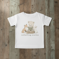 A white short-sleeve baby t-shirt with a cute graphic of a teddy bear, fox, and rabbit reading a book, with the text 'collecting stories' printed below. The t-shirt is laid flat on a rustic wooden plank background.