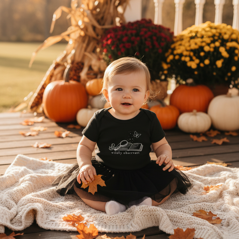 A baby sits on a white knit blanket on a wooden deck, wearing a black t-shirt with a white graphic of a pickup truck and the words 'Happy Autumn'. The background features autumn decorations including pumpkins, corn stalks, and yellow and red flowers.