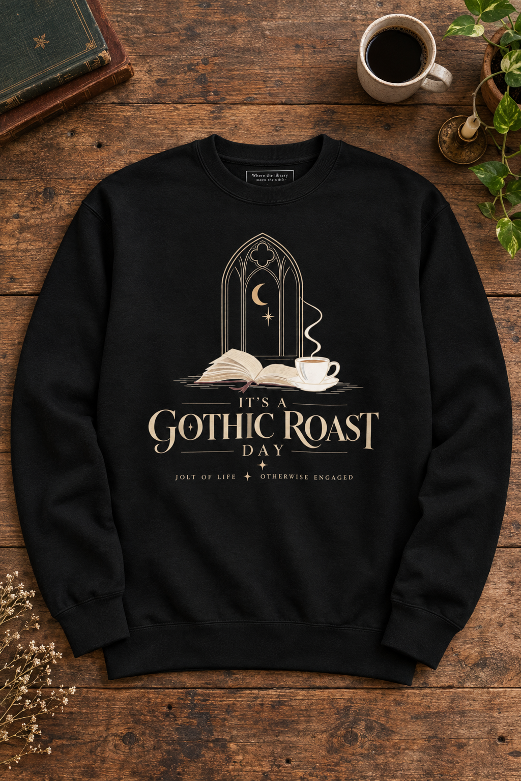 A black crewneck sweatshirt with a gothic-themed graphic print reading 'IT'S A GOTHIC ROAST DAY' and 'JOLT OF LIFE + OTHERWISE ENGAGED', laid flat on a rustic wooden table with a coffee cup, book, and plant nearby.