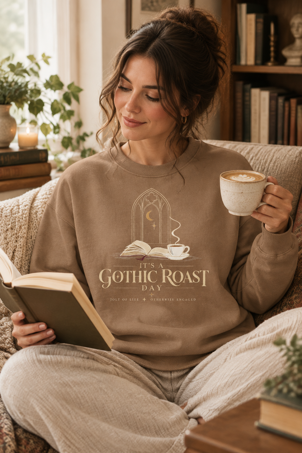 A woman sits on a couch in a cozy room, wearing a beige sweatshirt with a gothic-themed graphic that reads 'It's a Gothic Roast Day' and holding a mug while reading a book.