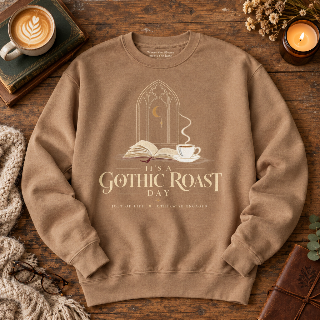 A tan crewneck sweatshirt laid flat on a rustic wooden table, featuring a gothic-themed graphic with an open book, a cup of coffee, and the text 'It's a Gothic Roast Day.' The sweatshirt is styled with cozy props including a cup of latte, a candle, glasses, a knit blanket, and a leather-bound book.