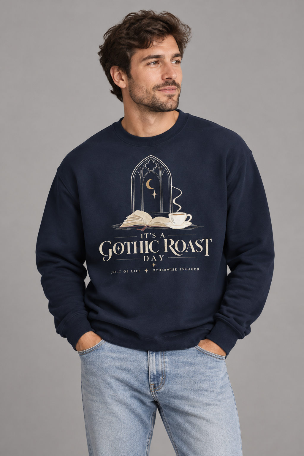 A man wearing a navy blue crewneck sweatshirt with a gothic-themed graphic that reads 'It's a Gothic Roast Day' and 'Jolt of Life • Otherwise Engaged' beneath it, paired with light blue jeans, against a solid gray studio background.