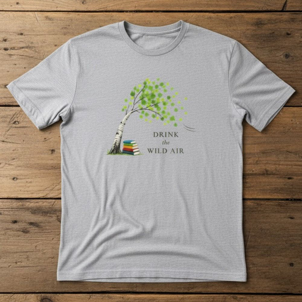 gray tee with 'drink the wild air' and tree illustration, blowing in the breeze, a stack of books rests under the tree