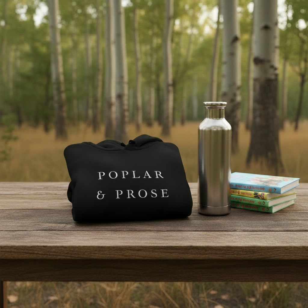 A black drawstring pouch with the words 'POPLAR & PROSE' in white serif lettering sits on a wooden table outdoors, next to a stainless steel water bottle and a stack of books, with a blurred birch forest background.