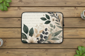 A laptop sleeve with a botanical floral pattern in green, beige, and white, laid flat on a rustic wooden surface with decorative leaves and cups at the corners.
