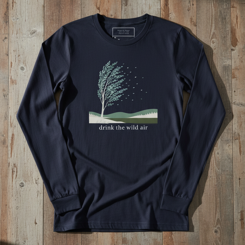 A navy blue long-sleeve t-shirt with a graphic of a tree on a hillside and the text 'drink the wild air' printed on the chest, laid flat on a rustic wooden plank background.