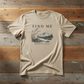 A beige crewneck t-shirt with a dark gray graphic print reading 'FIND ME WHERE IT'S QUIET' above a mountain and river landscape, laid flat on rustic wooden planks.