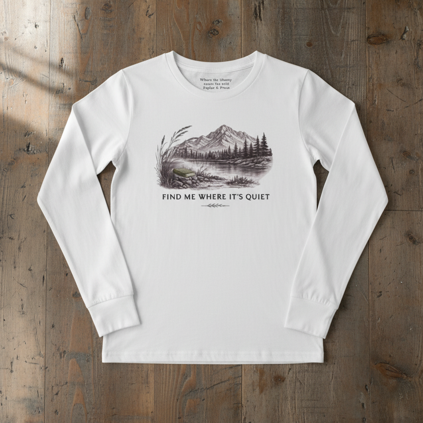 A white long-sleeve t-shirt with a black and white mountain lake graphic and the text 'FIND ME WHERE IT'S QUIET' printed on the chest, laid flat on a rustic wooden background.