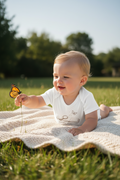 A baby lying on a white knit blanket in a grassy park, reaching toward a butterfly while wearing a white short-sleeve bodysuit with a butterfly graphic and the words 'BUTTERFLY ANNIVERSARY'.