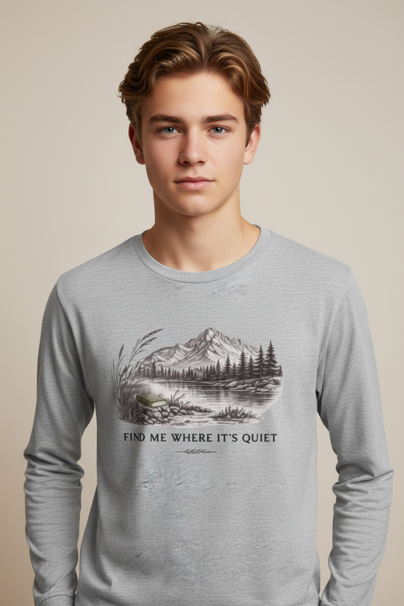 A young man wearing a light gray long-sleeve crewneck shirt with a black and white mountain lake graphic and the text 'FIND ME WHERE IT'S QUIET' on the front, set against a solid beige background.