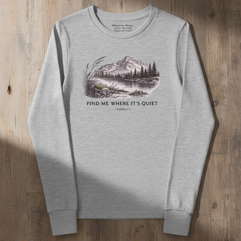 A light heather gray long-sleeve crewneck tee with a vintage-style mountain landscape graphic and the text 'FIND ME WHERE IT'S QUIET' on the front, laid flat on a rustic wooden background.