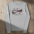 A light heather gray long-sleeve crewneck tee with a vintage-style mountain landscape graphic and the text 'FIND ME WHERE IT'S QUIET' on the front, laid flat on a rustic wooden background.